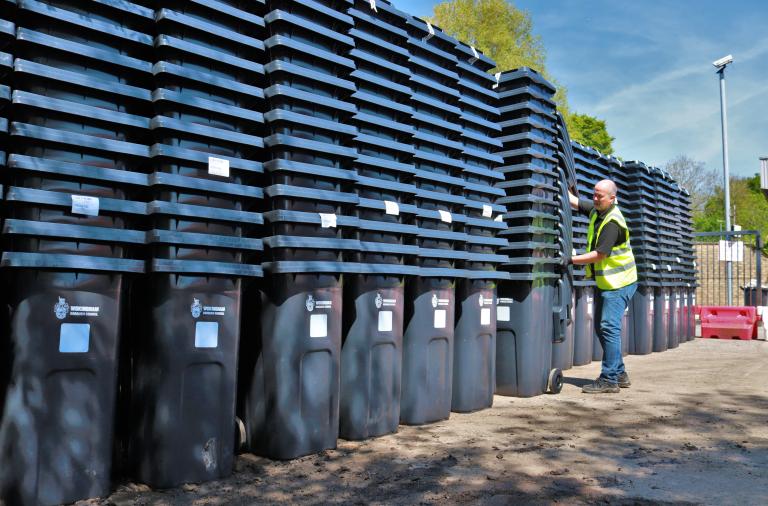 A man in a high vis jacket wheels a tall stack of bins up against many other tall stacks of bins in a storage yard