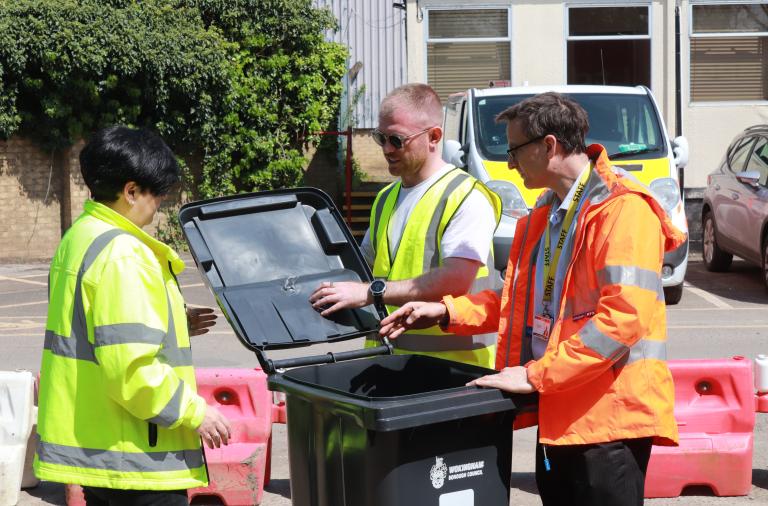 A man in a high vis jacket demonstrates a new bin to two other people in high vis jackets