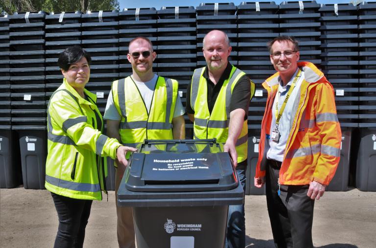 Four people in high vis jackets gather around a new bin and smile to the camera, with several huge stacks of new bins behind them