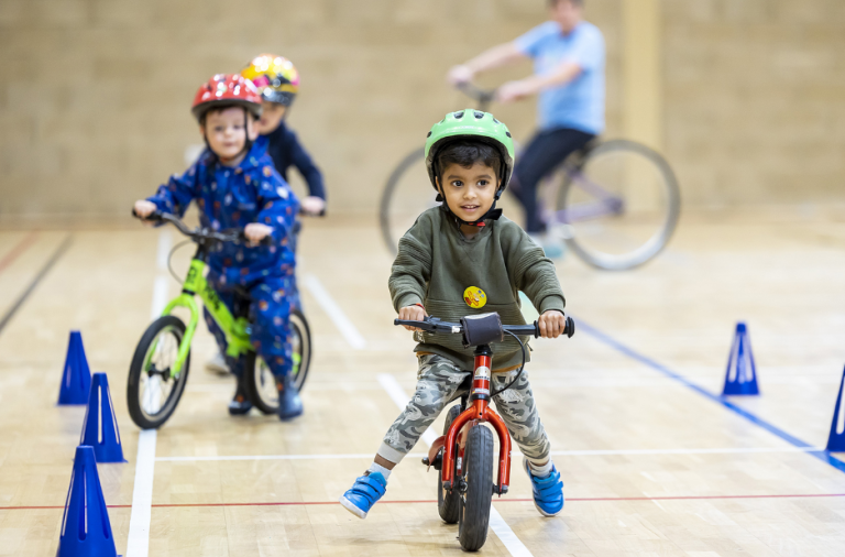 A young child in a cycle helmet rides a balance bike between some cones during a group session
