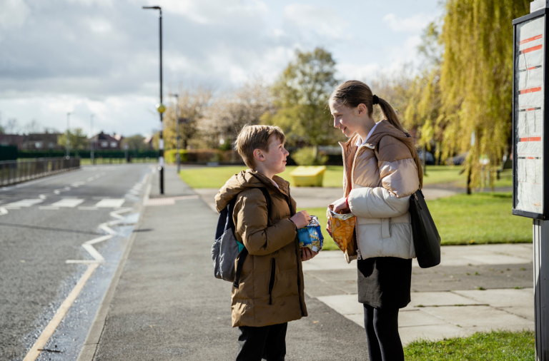A boy and girl eating bags of crisps stood at a bus stop