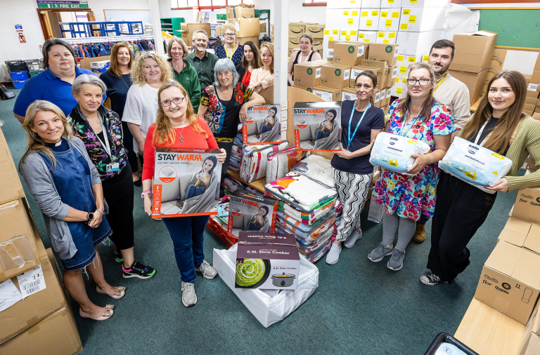 17 people from winter warmers network smile to the camera surrounded by boxes, some are holding energy efficient items