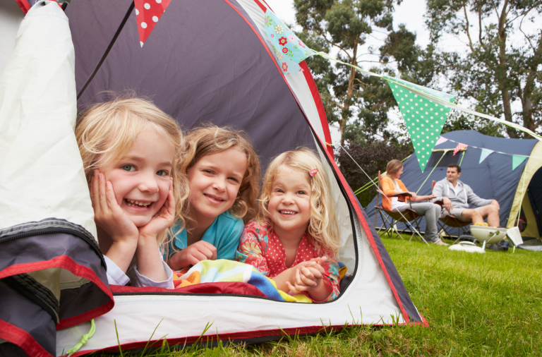 Three girls inside a tent smile to the camera through the hatch opening, bunting is decorating the area around their tent