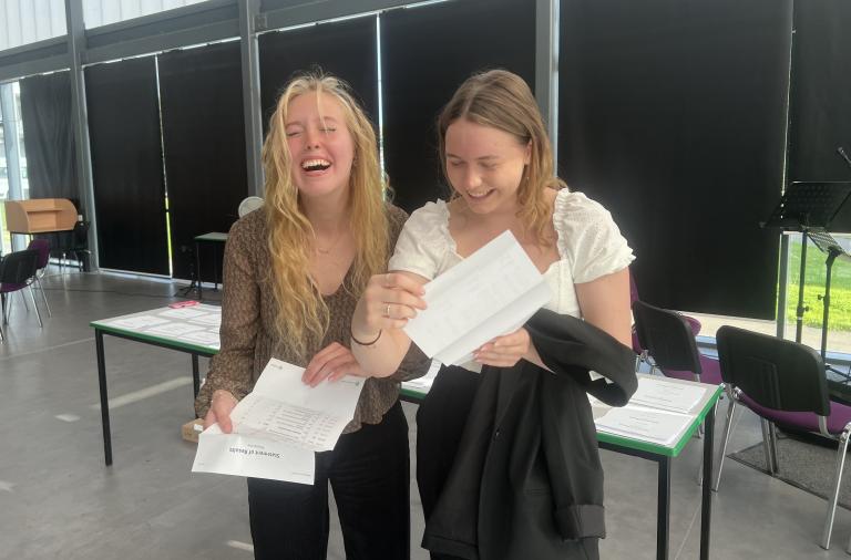 Two female students smile and laugh as they look at their exam results
