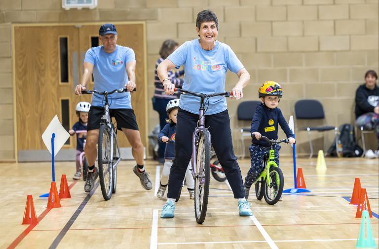 A woman guides a bicycle along a track of cones in a sports hall, followed by other adults and children