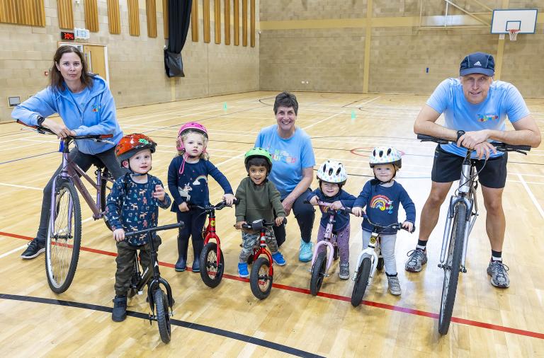 A group of adults and very small children, on bikes of different sizes, smile for the camera inside a large sports hall