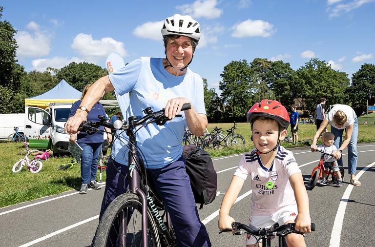A woman in a council T-shirt smiles and poses on a bike next to a young girl, also on a bike, on an outdoor track on a sunny day