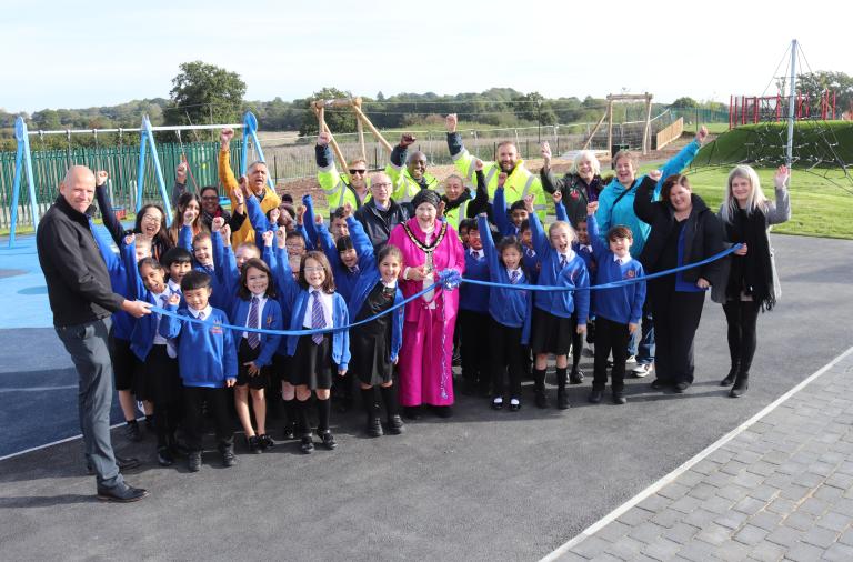 Children and adults cheer as the Mayor cuts a blue ribbon to open a playground, seen behind the group