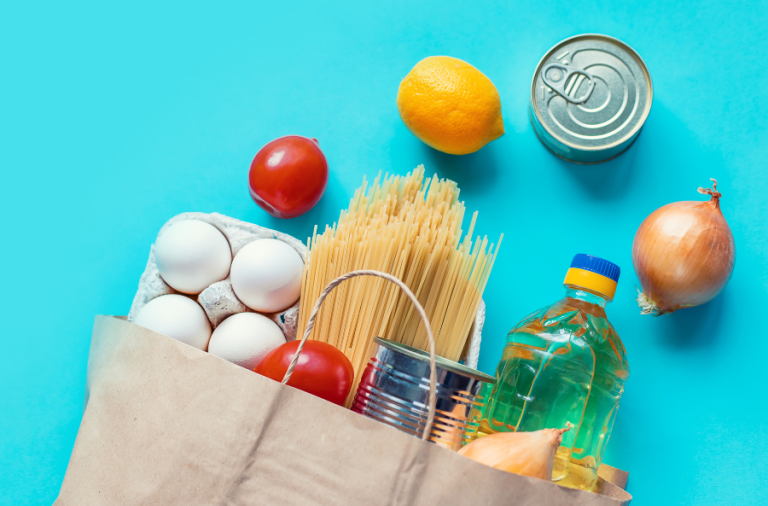 A paper bag of groceries on it's side with eggs, spaghetti, onion, tomatoes, a lemon, a bottle of oil and two tins spilling out