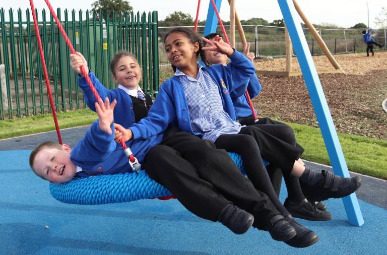 Four children crowd on to a swing with a large seat and wave at the camera, with one almost hanging upside down 