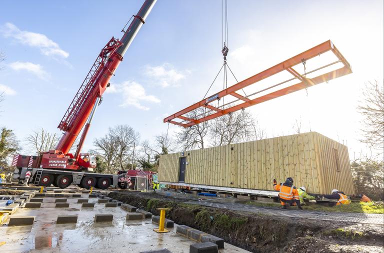 Photograph of a crane lifting the roof onto a rectangular, single-storey timber-clad building on a building site