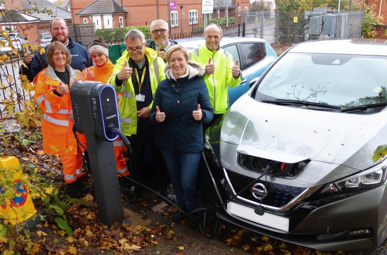 A group of men and women, many in high vis gear, cheer and give the thumbs up next to two cars charging side by side in a car park