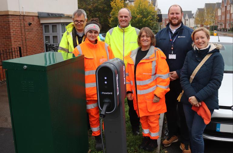 A group of men and women, many in high vis gear, stand next to a chargepoint and power cabinet on a residential street