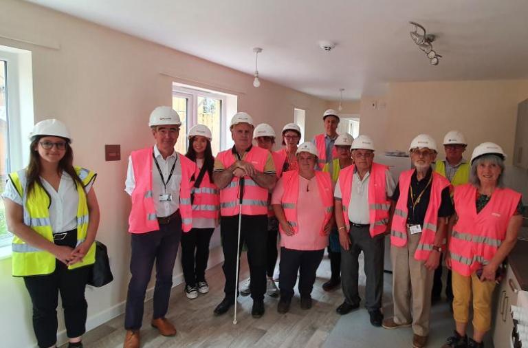 A group of adults wearing high viz jackets in pink and yellow and wearing hard hats stands inside one of the new modular homes at Grovelands
