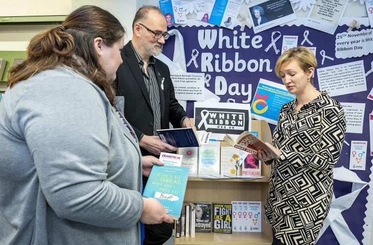 Three adults stand by a stand containing white ribbon related books in the library against a backdrop of a white ribbon day display 