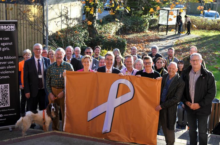 Councillors and officers stand outside the council building holding up the white ribbon flag, an orange flag with a white ribbon, next to a pull up banner with information about white ribbon