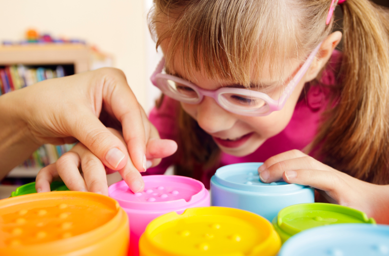 A girl leans over a set of textured stacking cups, with an adult helping her to feel the surface