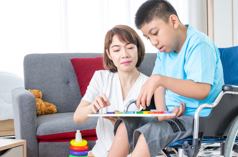 A boy sits in a wheelchair with an adult beside him holding an insert jigsaw