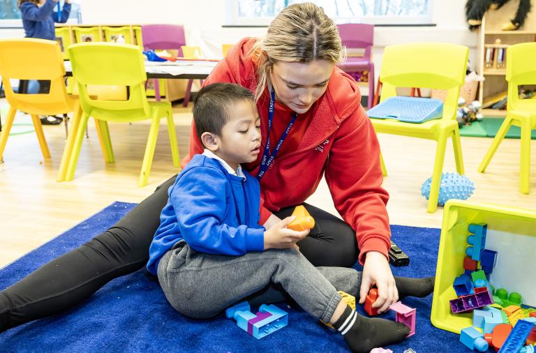 A child and school staff sit on the floor playing with bricks