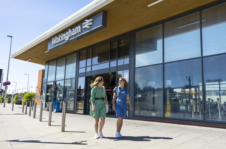 Two women smile and chat with each other as they walk past Wokingham railway station on a summer day