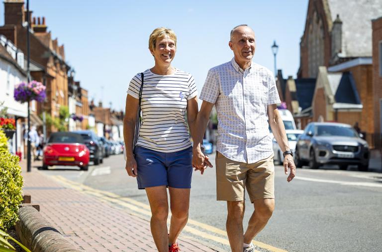 An older couple walk together hand in hand along a pavement in a town centre on a bright, sunny day