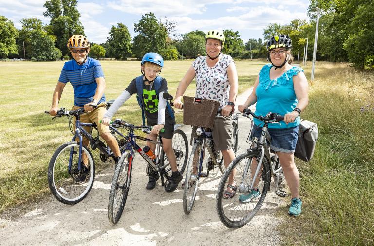 A family pose for the camera while sitting on bikes and wearing helmets. They have stopped on a cycle way running through long, tall grass on a summer day.