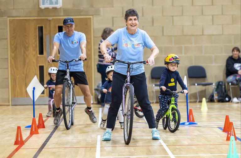 Photo of Jill Bissell riding a bike around an obstacle course in a sports hall as little children watch and learn