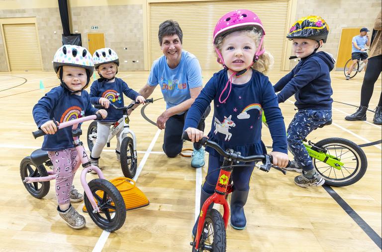 A group of small children in helmets, including one looking right at the camera, gather around Jill in a sports hall