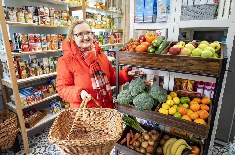 Cllr Rachel Bishop-Firth with a shopping basket at Roots Community Store, next to a shelf of fruit and vegetables, with other goods on show behind her