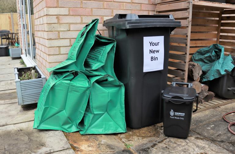Four green recycling bags, a black wheeled rubbish bin marked "your new bin" and a food waste bin behind a house