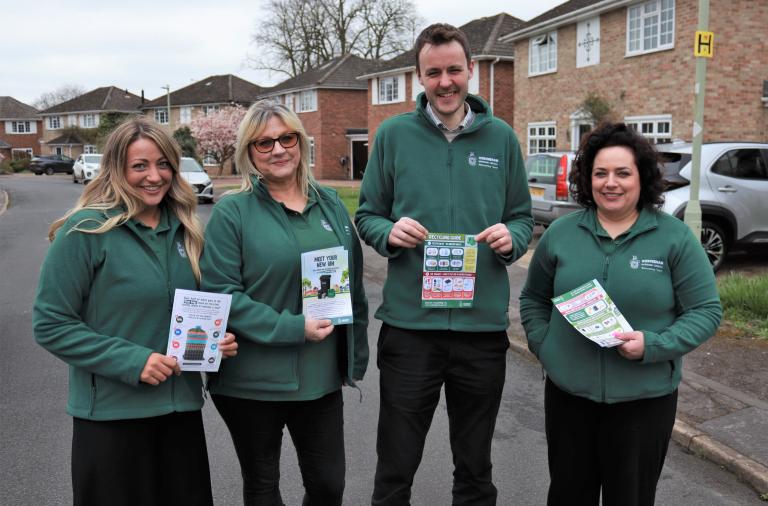 Three women and a man wearing dark green council jackets, standing in a residential street and holding up information leaflets
