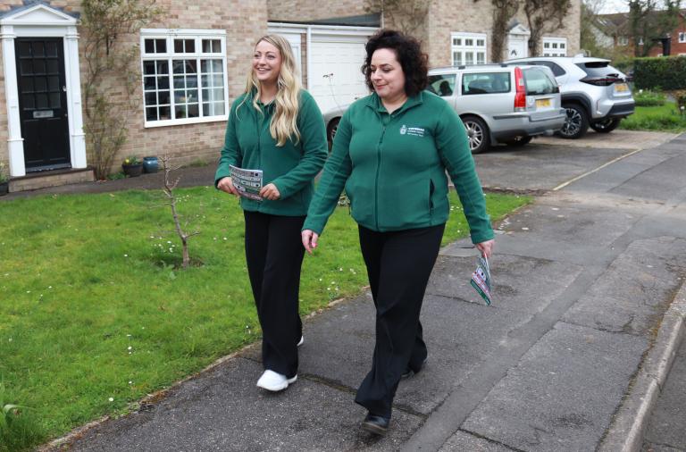 Two women in dark green council jackets smile and chat with each other as they walk down a street holding leaflets
