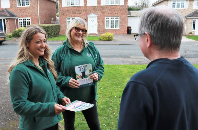 Two smiling women in green council jackets chat with a man on his doorstep while pointing to an information leaflet about waste collections