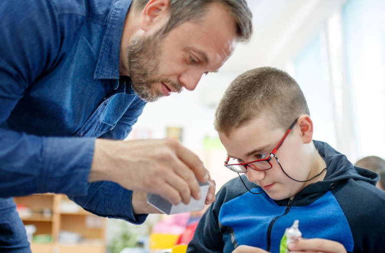 A boy is in a classroom working on a piece of craft with the help of an adult male