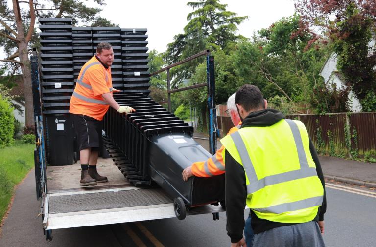 Two delivery men in high vis jackets unload a large stack of bins from the back of a small lorry