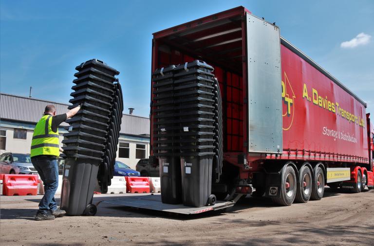 A man in a high vis jacket unloads several stacks of black bins from a big lorry