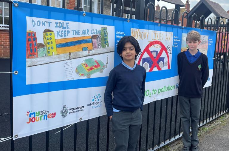 Two boys stand smiling in front of a large banner at the school gates displaying their artwork
