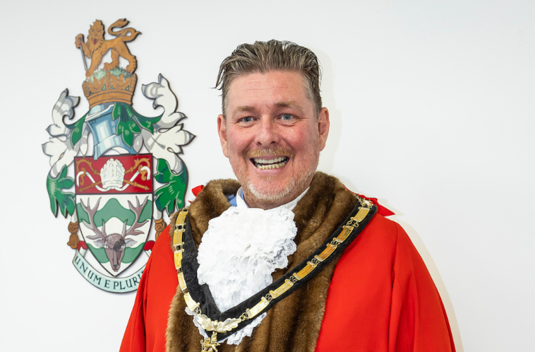 Cllr Adrian Mather in his mayoral regalia and chain, next to the council's emblem