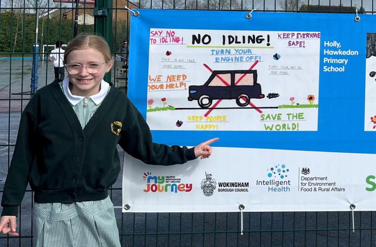 A smiling girl points at her no idling artwork, which is printed on a large banner