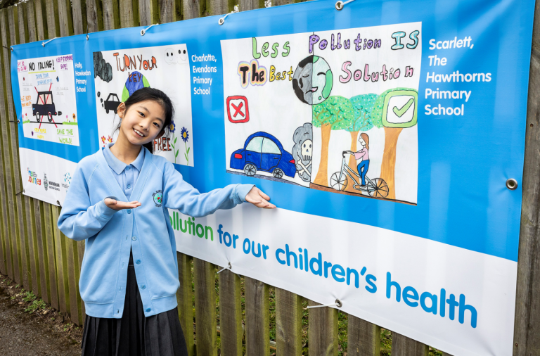 A young girl beams proudly next to her artwork which is printed on a big banner at her school