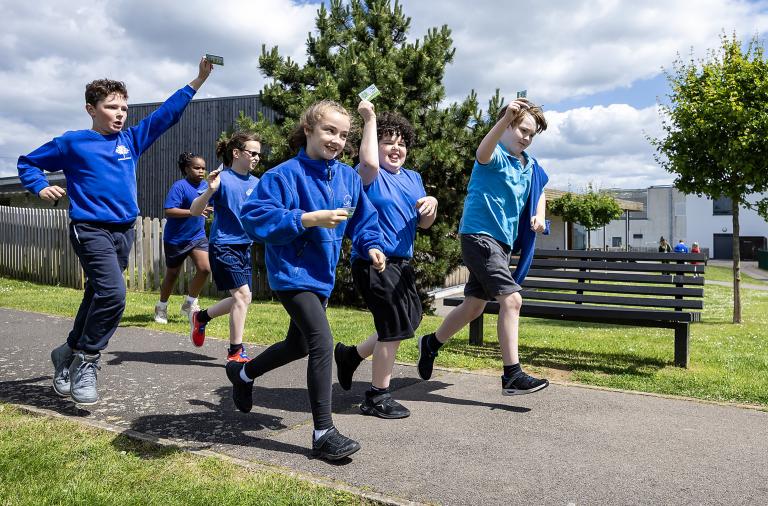 A group of excited children rush along a path holding up contactless score cards