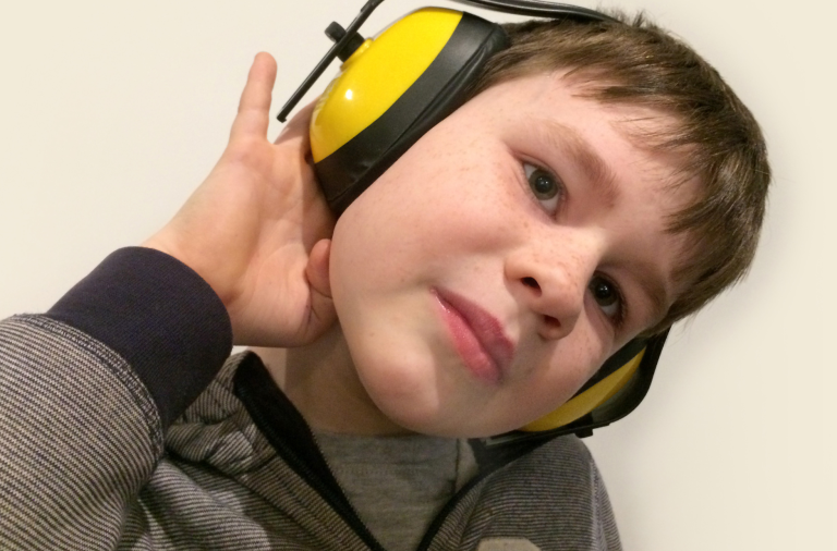A boy wearing yellow ear defenders tilts his head on one side while looking at the camera