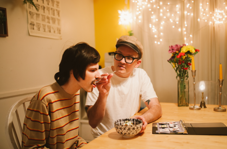 A man supports another man with eating from a bowl at a table