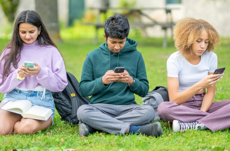 Three teenagers sitting cross legged on the grass looking at their mobile phones