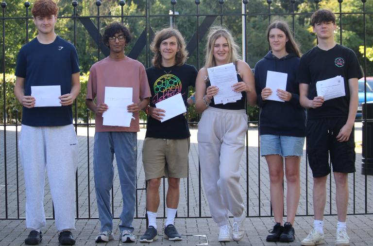 Six students stand in a row smiling and holding up their A level results
