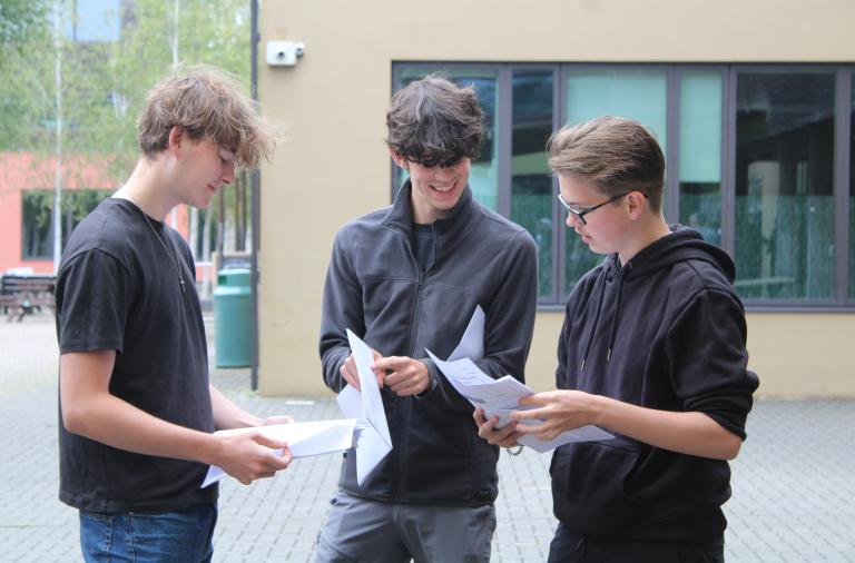 Three students stand together smiling and sharing their grades with each other