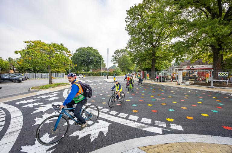 A group of adult and child cyclists ride across a new junction at California Cross with colourful leaf patterns on the surface