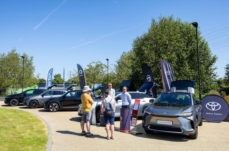 A line of electric vehicles parked outside a business park, with members of the public looking at them