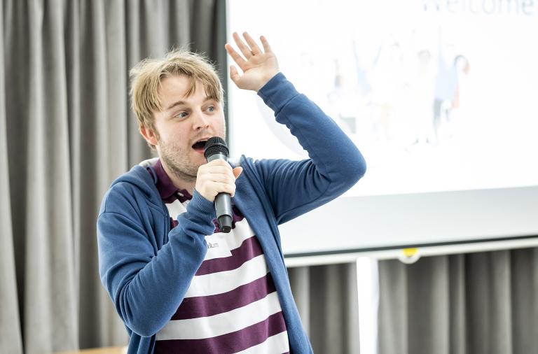 A man stood in front of a presentation speaks into a microphone