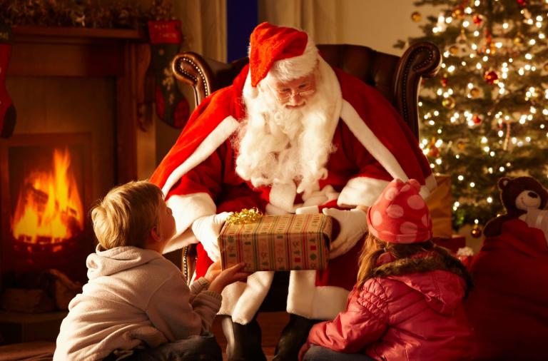 Santa with two children giving a gift. He is next to a Christmas tree. 
